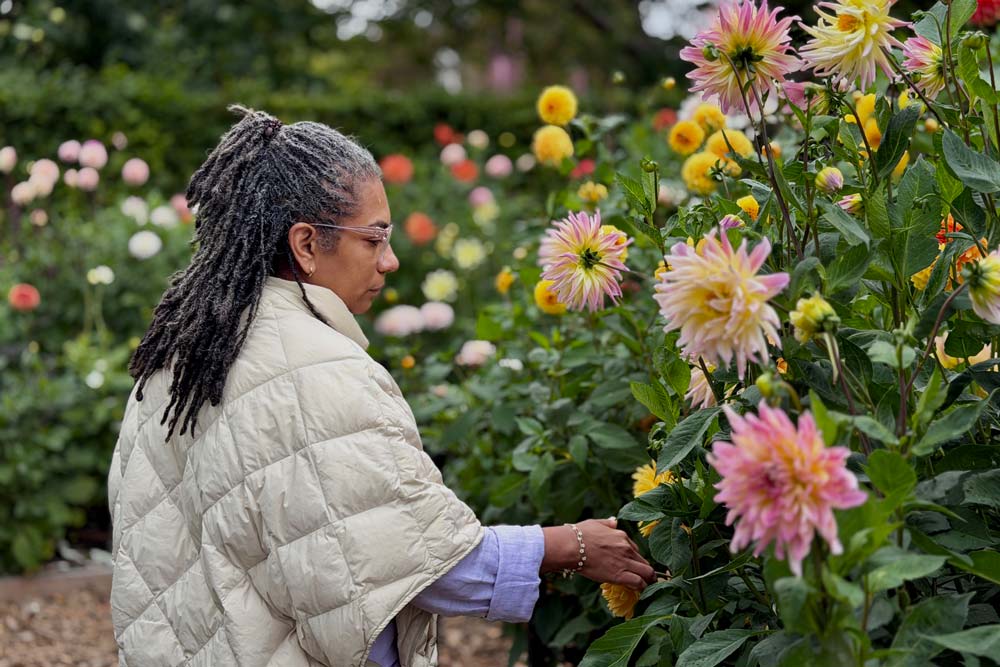 Kvinde med grå dreadlocks rører ved en dahlia i haven — selvpleje mellem kropsterapi-behandlinger i Højbjerg